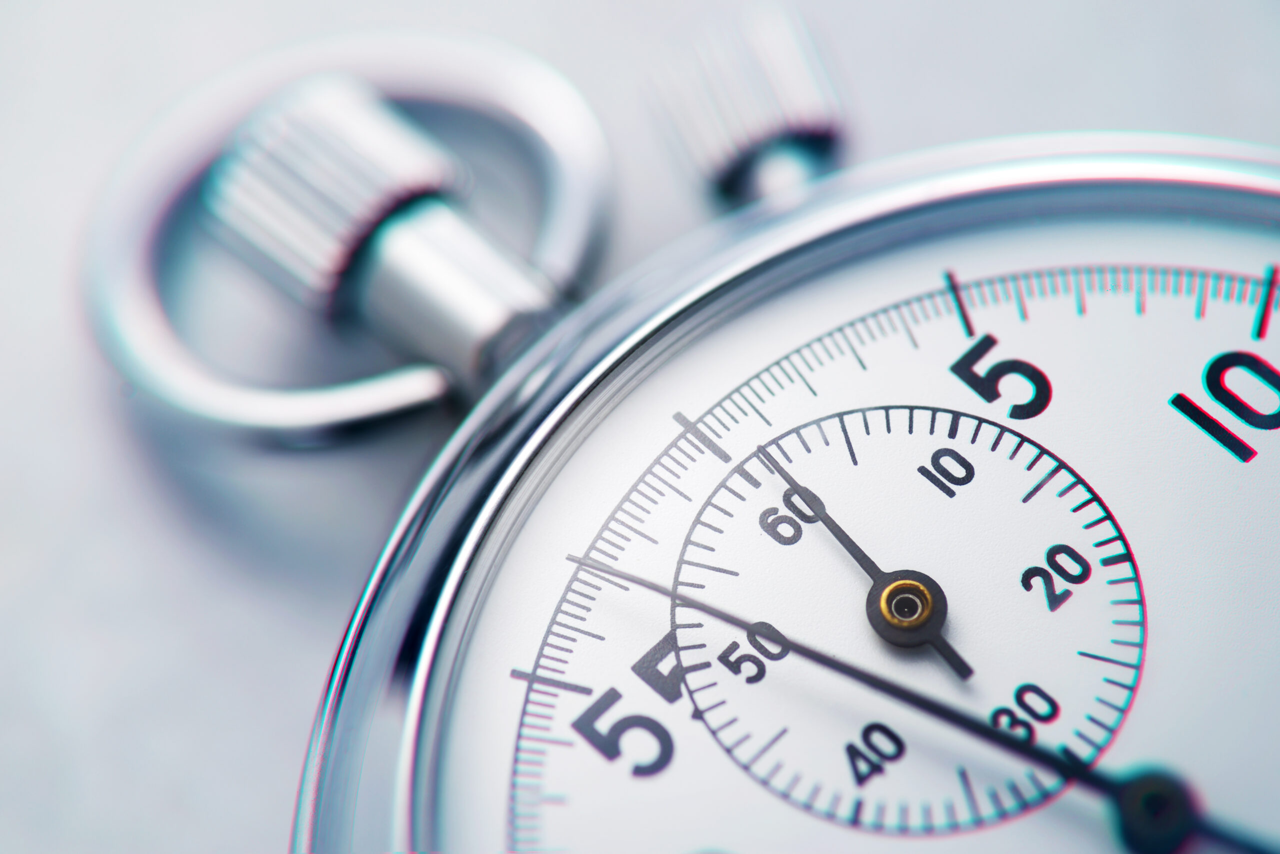 Close-up macro photograph of a chrome stopwatch on a pale background, second hand pointing near the 60 mark, with a slight chromatic-aberration color shift on the dial markings.